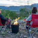 The Live Outdoor Firestorm Series I Portable Propane Fire Pit is being used outdoors, providing a great view of the mountains, with people sitting nearby.