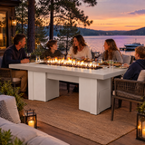 Family dinner on a lakeside deck at twilight around a Top Fires Alameda 72-inch GFRC gas fire table with a Limestone tabletop and White base