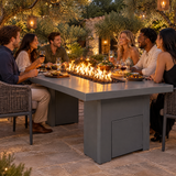 Friends sharing a meal in a garden courtyard with olive trees around a Top Fires Alameda 72-inch GFRC gas fire table with a Natural Gray tabletop and Gray base
